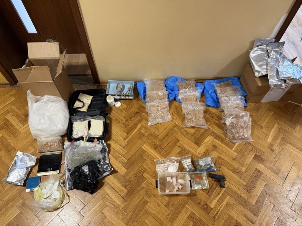 Na podłodze w pomieszczeniu znajdują się różne pakunki, w tym torby z substancjami, waga i pistolet.