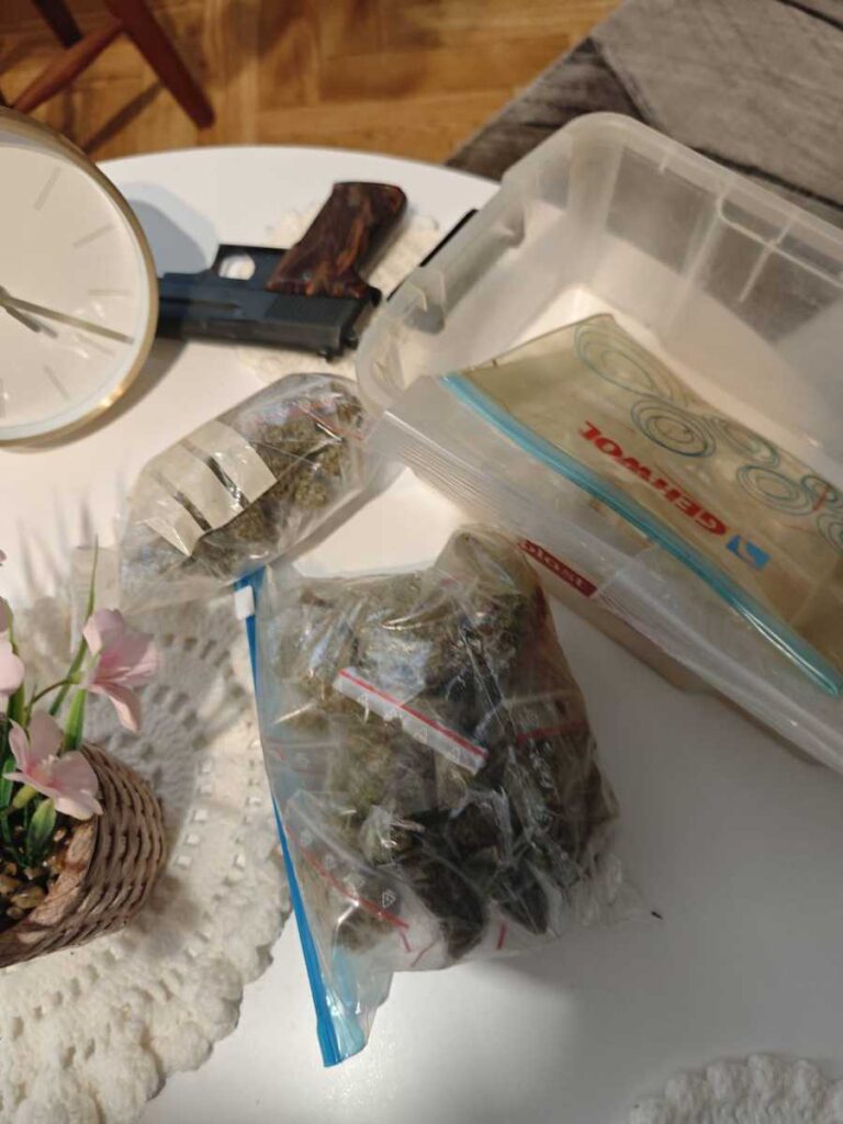 Na stoliku znajduje się pistolet, woreczki z substancją roślinną oraz zegar. Obok leży ozdobna doniczka z kwiatami.