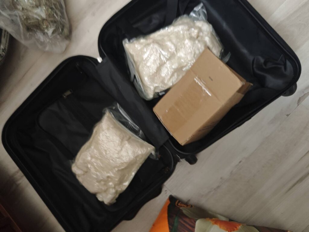 Walizka z pakunkami wypełnionymi foliowymi torbami oraz kartonem, leżąca na podłodze w pomieszczeniu.