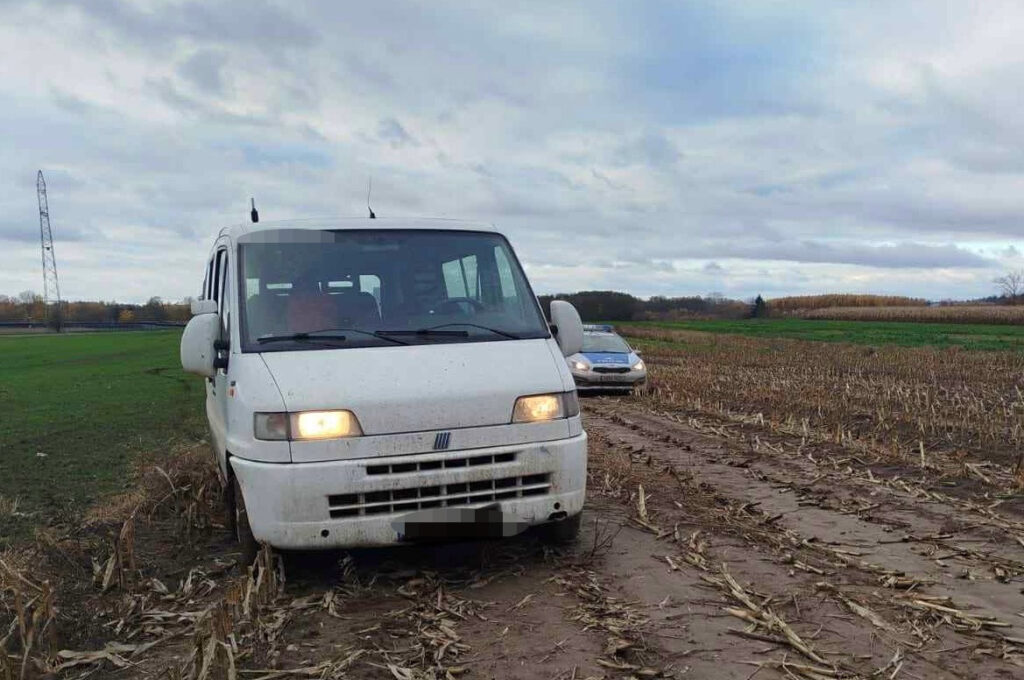 Biały bus utknął w błotnistej drodze na polu, obok stoi samochód osobowy. W tle widoczne są pola i chmury.