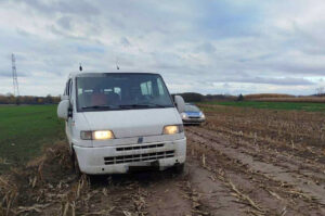 Biały bus utknął w błotnistej drodze na polu, obok stoi samochód osobowy. W tle widoczne są pola i chmury.