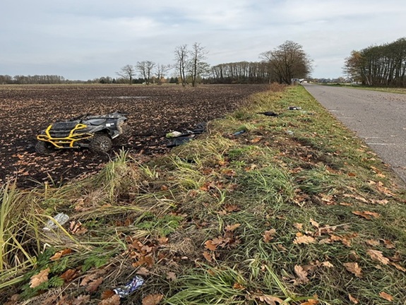 Na polu widać uszkodzony quad oraz porozrzucane przedmioty, obok znajduje się asfaltowa droga i zadrzewienie w tle.