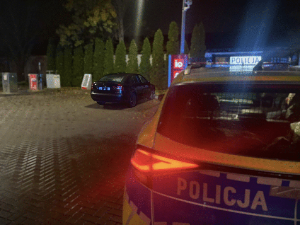 Czarny samochód osobowy stoi na parkingu obok stacji paliw, w tle widoczny radiowóz policji z włączonymi sygnałami.