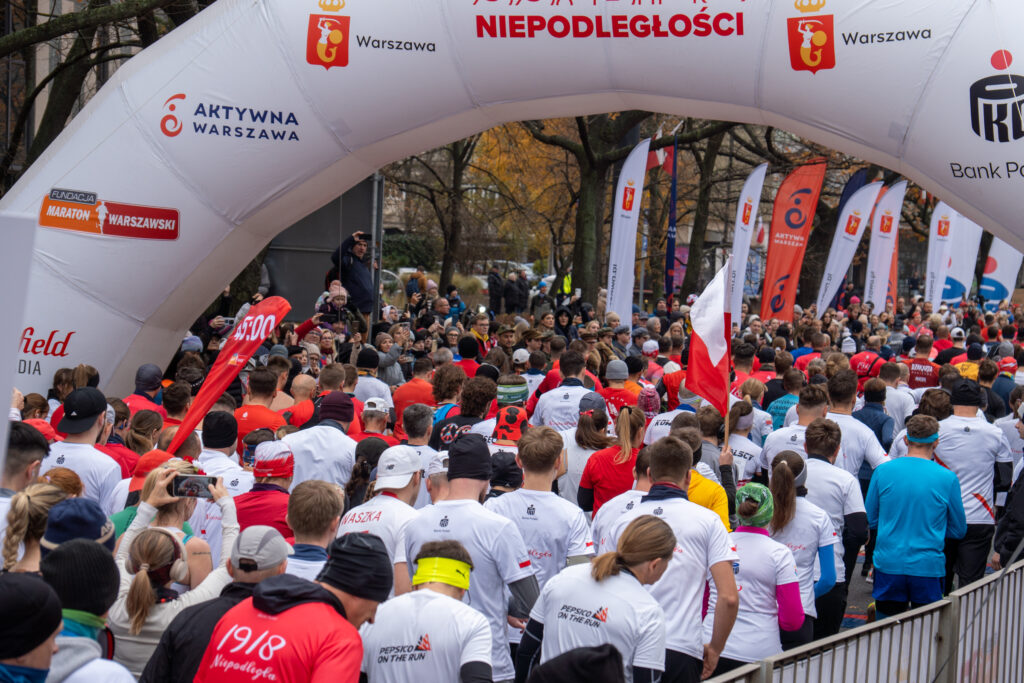 Biegacze w białych i czerwonych koszulkach startują w wydarzeniu biegowym pod łukiem startowym z logo miasta.