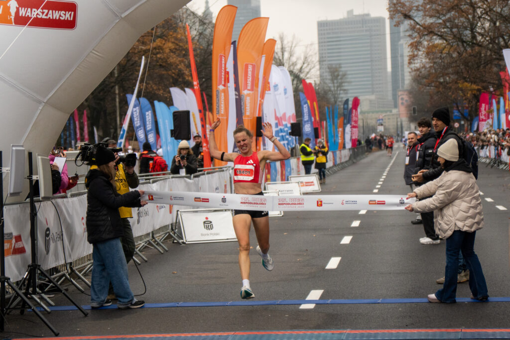 Biegaczka w czerwonym stroju przekracza metę, unosząc ręce w geście radości na ulicy z transparentami w tle.