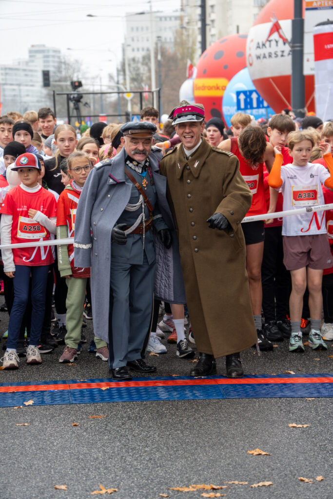 Dwaj mężczyźni w historycznych mundurach pozują do zdjęcia na tle grupy dzieci i młodzieży przed startem biegu.