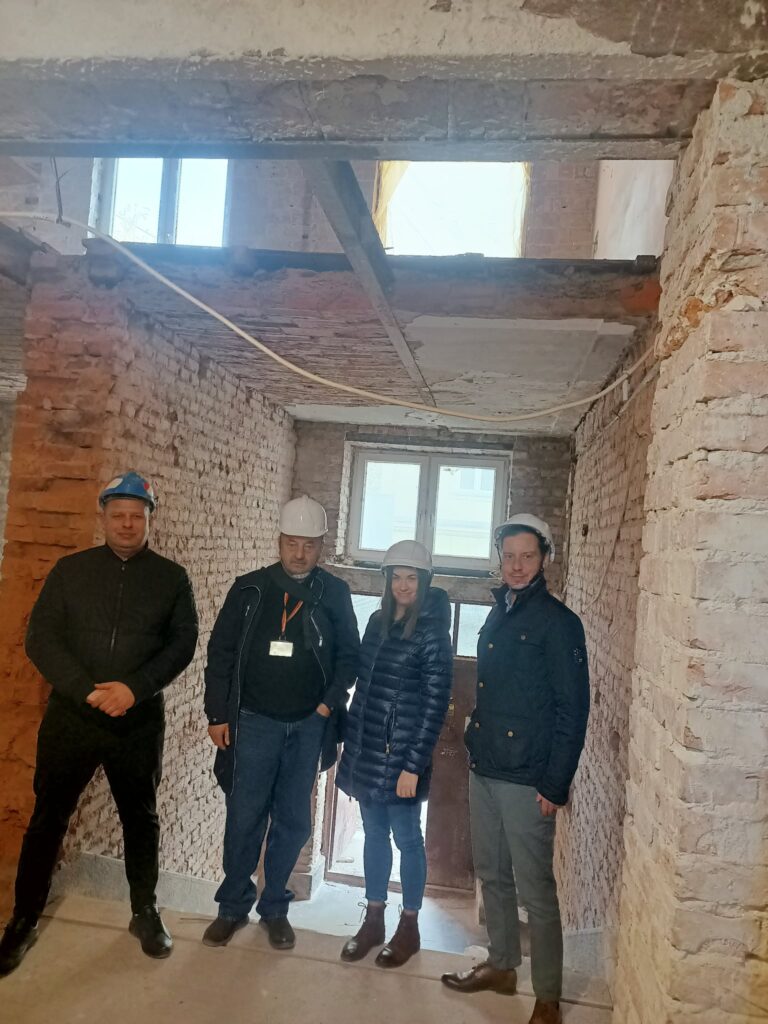 Czterech pracowników w kaskach stojących w pustym wnętrzu budynku z ceglaną ścianą i oknami.