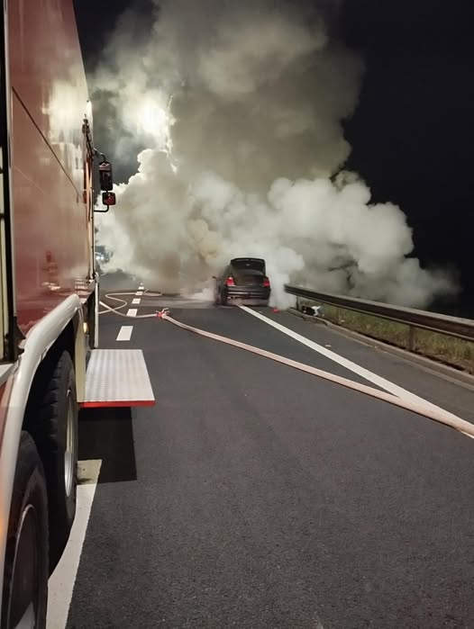 Samochód unosi się w chmurze dymu na drodze, obok węże strażackie, w tle widoczny wóz strażacki.