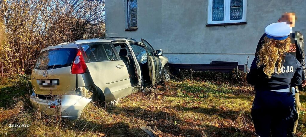 Uszkodzony samochód osobowy stoi w zaroślach obok budynku, a policjantka rozmawia z mężczyzną w pobliżu.