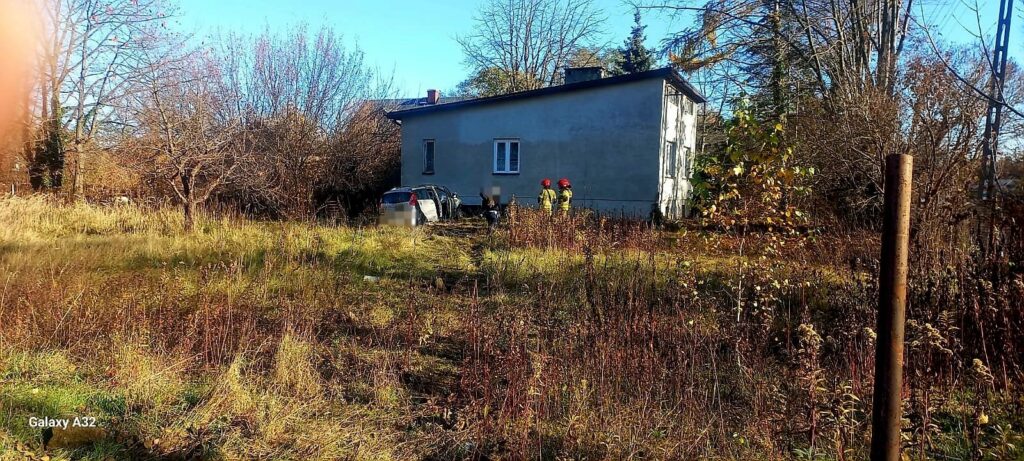 Grupa osób w odblaskowych strojach z czerwonymi kaskami przebywa w zarośniętym terenie obok szarego budynku.