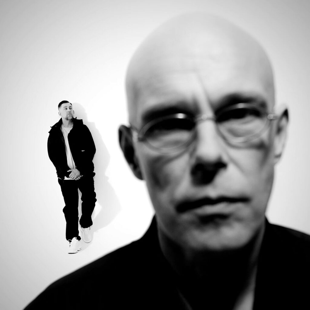 Mężczyzna z okularami w zbliżeniu, a w tle stoi inny mężczyzna w ciemnym ubraniu.