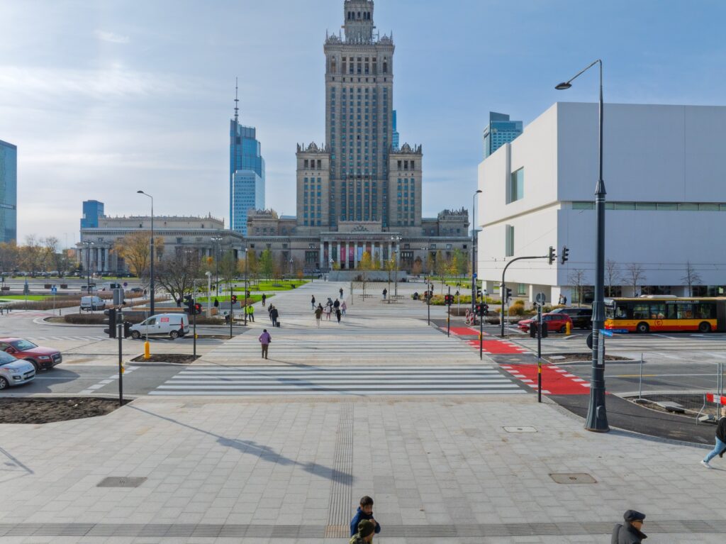 Wielu przechodniów spaceruje po nowoczesnym placu z widokiem na wysoki, zabytkowy budynek oraz nowoczesne wieżowce.
