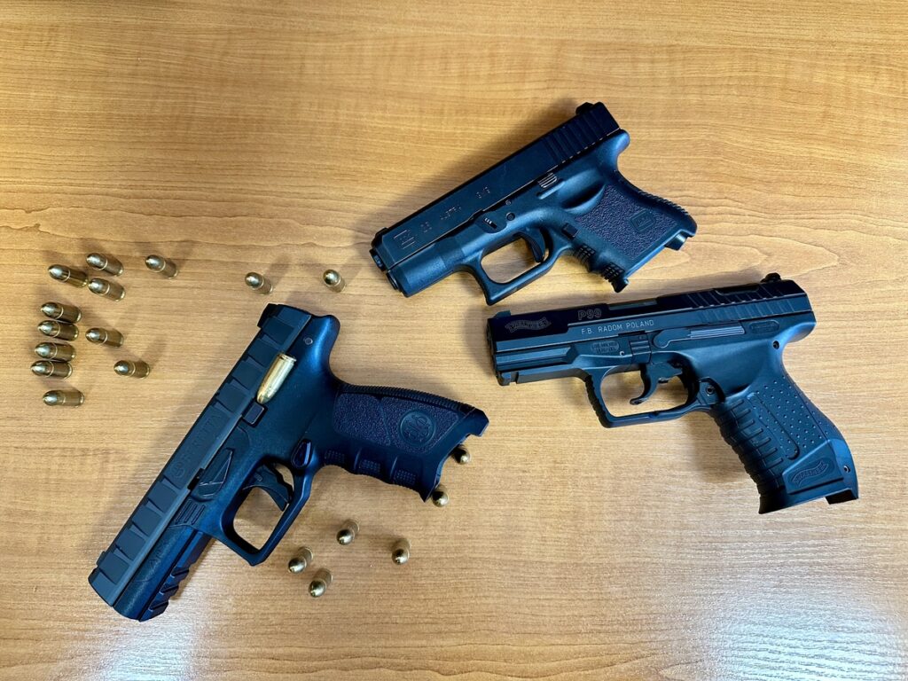 Trzy czarne pistolety leżą na drewnianym stole obok złotych nabojów.