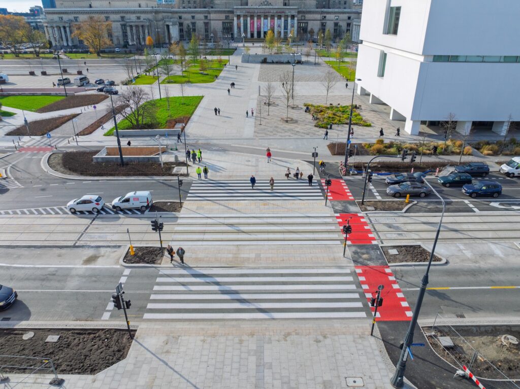 Grupa ludzi przechodzi przez pasy, obok nowoczesnego budynku i ruchliwej ulicy z samochodami.