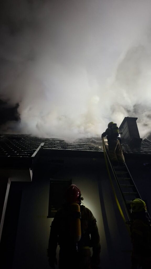 Strażak w hełmie wspina się po drabinie na dach budynku, z którego wydobywa się gęsty dym.