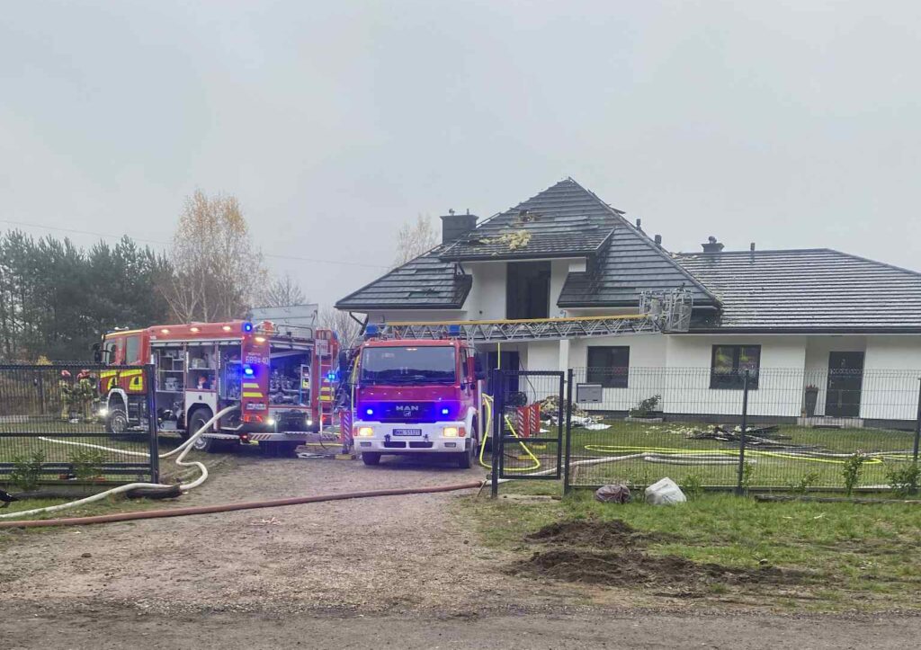 Dwa wozy strażackie z włączonymi sygnalizacjami stoją przed nowoczesnym domem z uszkodzoną dachówką.