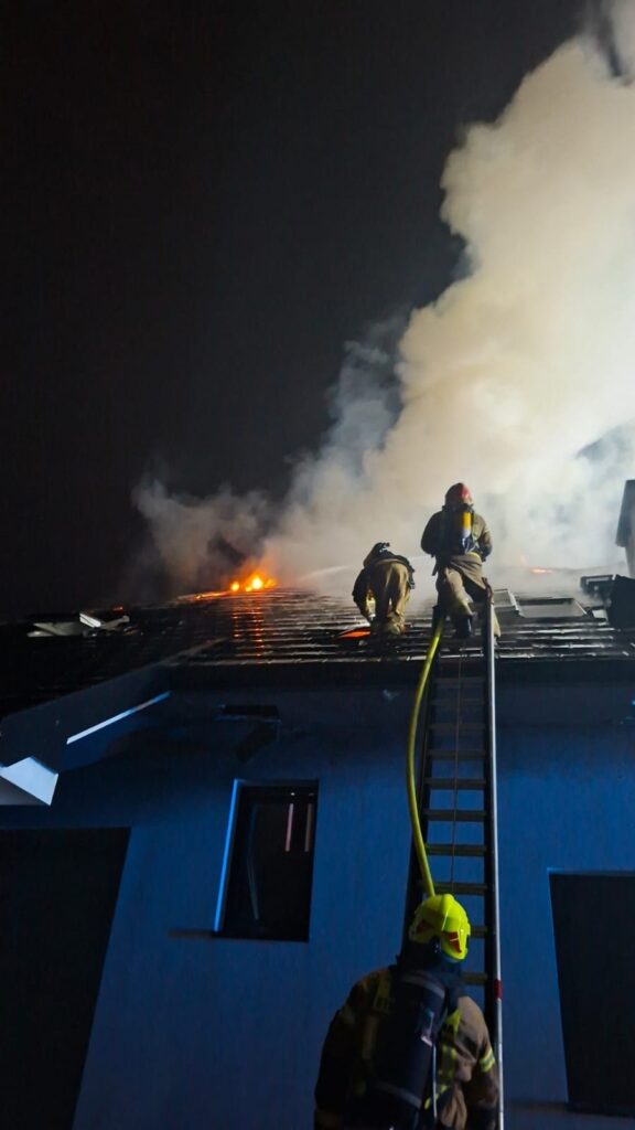 Strażacy na dachu budynku gaszą płomienie, otoczeni gęstym dymem w nocy.