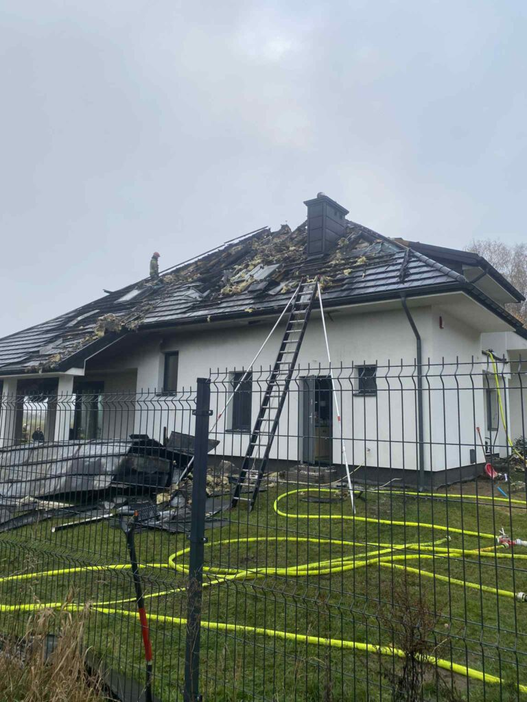 Strażak na dachu uszkodzonego domu, obok drabina oraz węże strażackie na trawie.