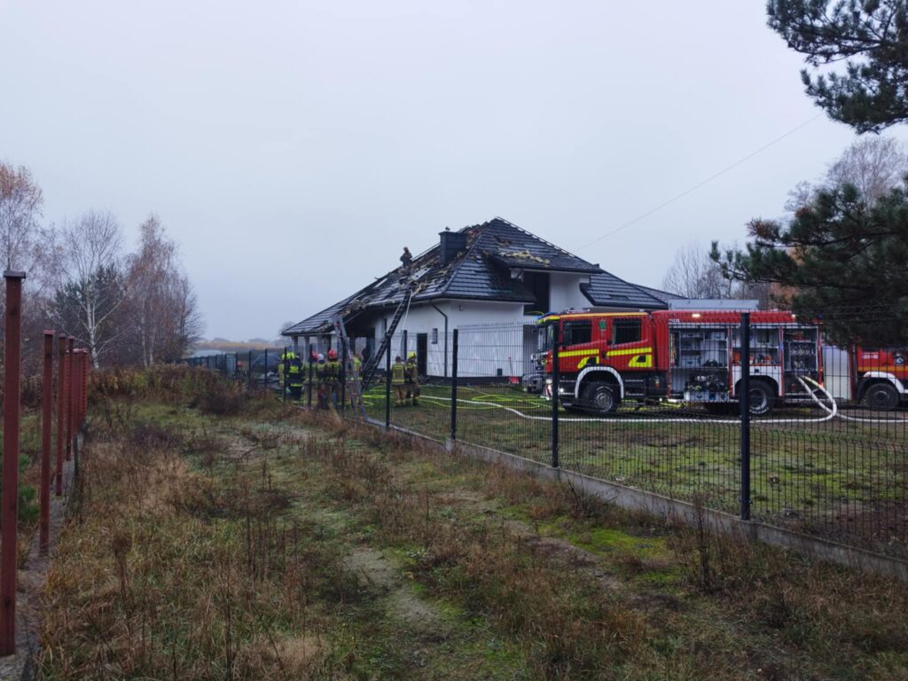 Strażacy pracują nad dachem uszkodzonego budynku mieszkalnego, obok stoi wóz strażacki. W tle widoczna siatka ogrodzeniowa.