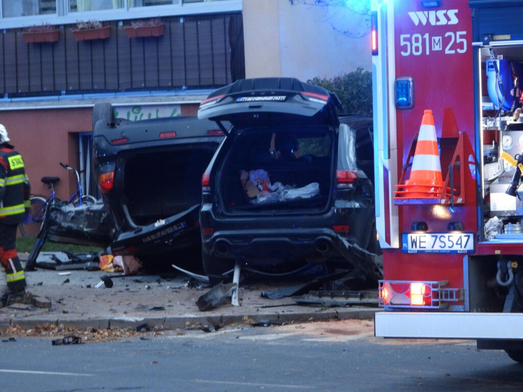 W miejscu wypadku leżą przewrócone samochody, a obok nich strażak w mundurze, przy włączonym wozie strażackim.