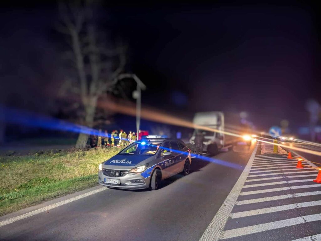 Policyjny radiowóz z włączonymi sygnałami świetlnymi stoi przy drodze, obok grupy ratowników i ciężarówki.