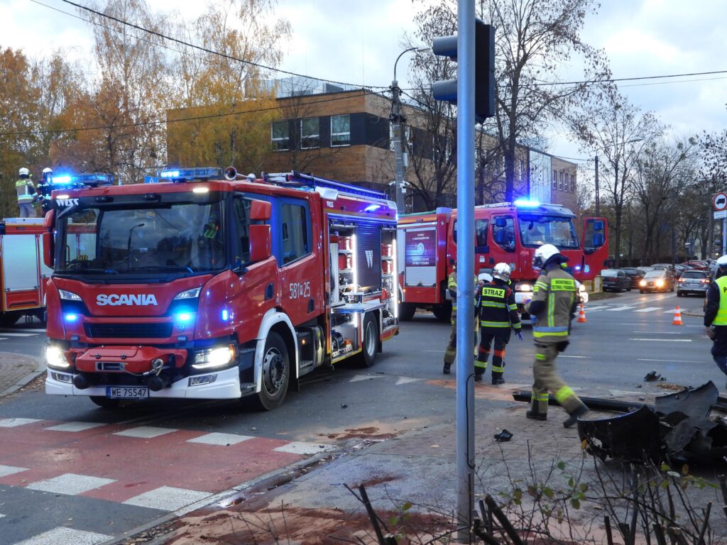 Czerwony wóz strażacki z włączonymi sygnałami świetlnymi stoi na skrzyżowaniu, a strażacy w mundurach badają miejsce zdarzenia.