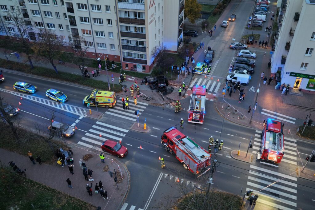 Na skrzyżowaniu ulicy stoją radiowozy, karetka oraz wozy strażackie, a obok zbierają się gapie.