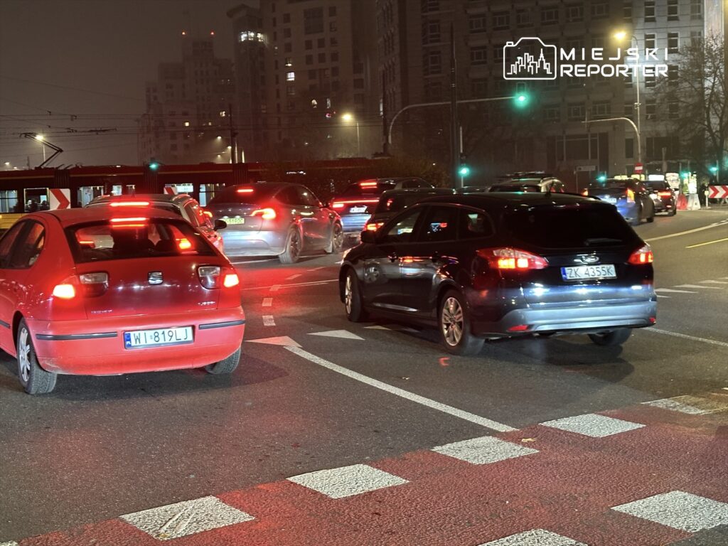 Ruch samochodowy na skrzyżowaniu w nocy, z widocznymi czerwonymi światłami hamowania i migającymi sygnalizatorami.