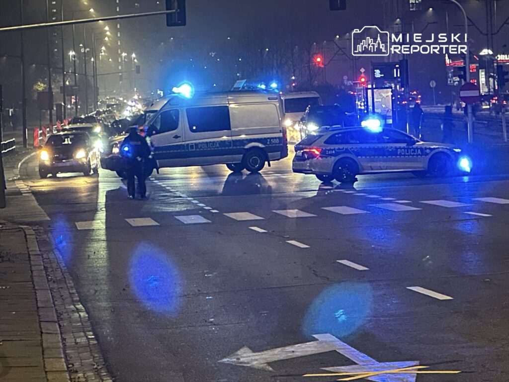 Policyjni funkcjonariusze kierują ruchem na zablokowanej ulicy, obok zaparkowanych radiowozów z włączonymi sygnałami świetlnymi.