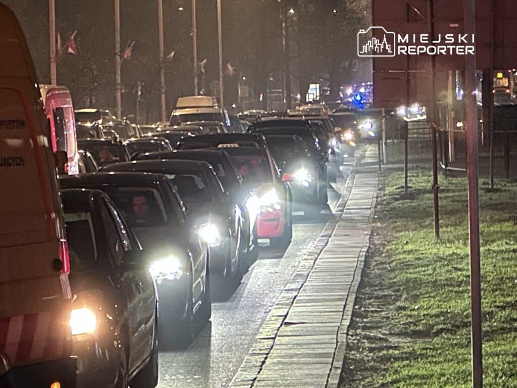 Długi korek samochodowy na ulicy, z pojazdami stojącymi w nocy, oświetlonymi przez reflektory.