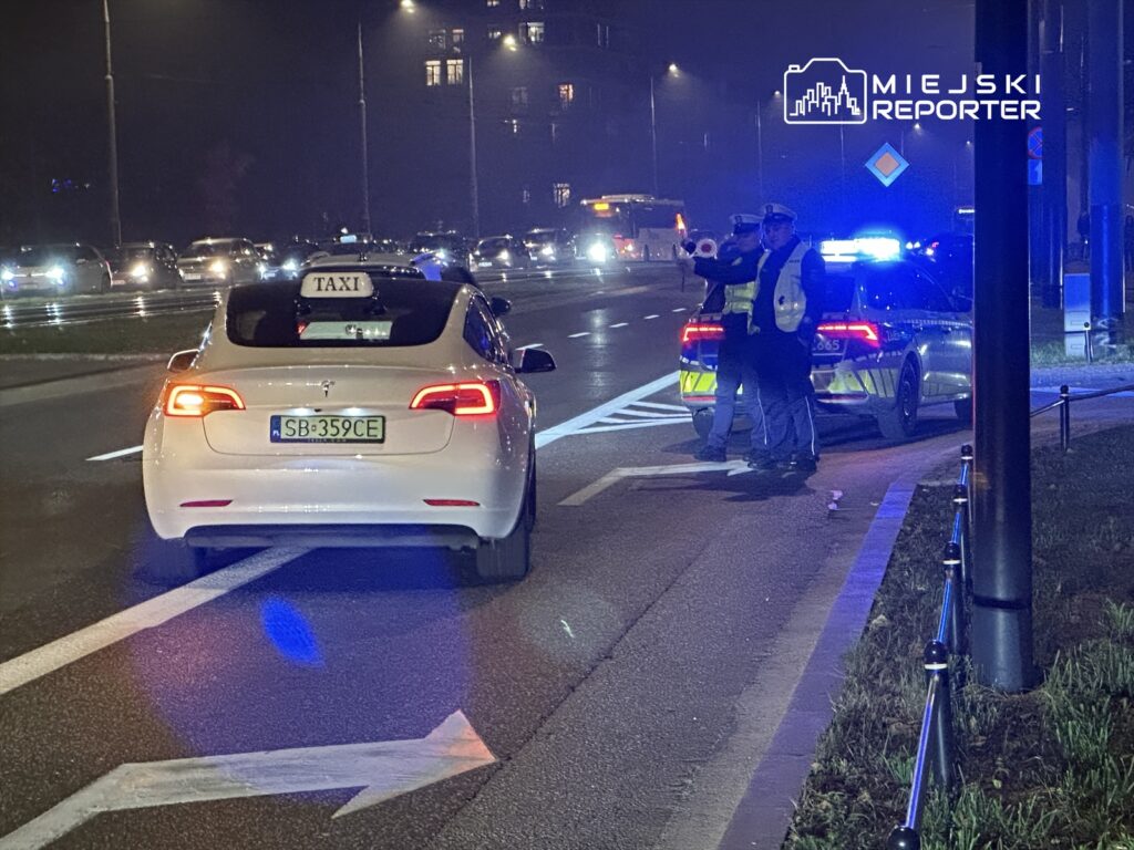 Biały samochód osobowy z oznaczeniem "TAXI" zatrzymany przez policjantów na drodze w nocy.