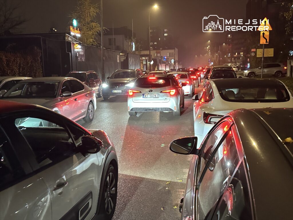 Ruch samochodowy w godzinach wieczornych na oświetlonej ulicy, z wieloma pojazdami stojącymi w korku.