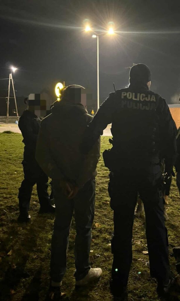 Policjant w mundurze zatrzymuje mężczyznę z rękami za plecami na oświetlonym terenie.