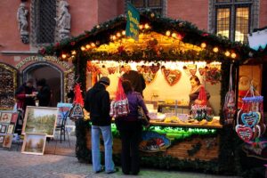 Kupujący z torbami stoją przy straganie ze słodyczami, w tle widoczne obrazy na sztalugach oraz dekoracje świąteczne.