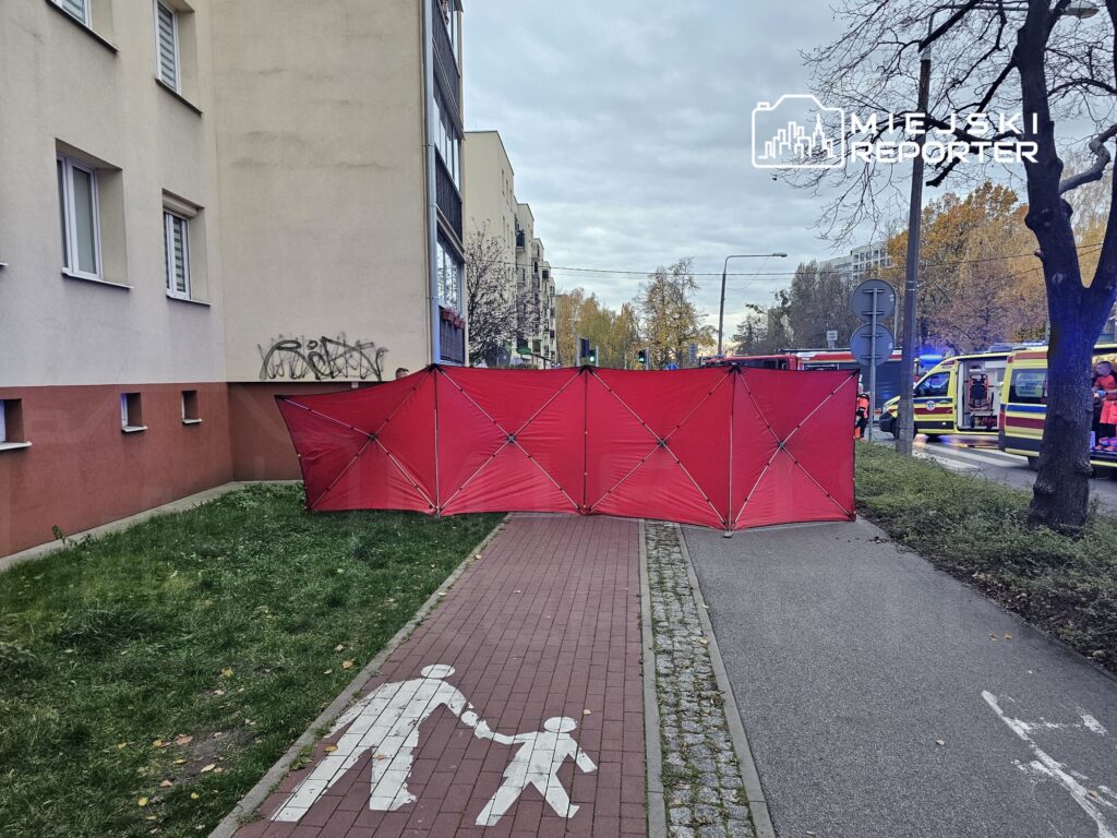 Czerwony namiot zabezpieczający ustawiony przy chodniku obok budynku, w tle widać pojazdy służb ratunkowych.