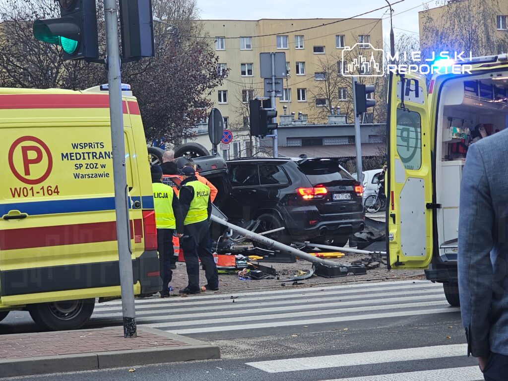 Na ulicy, w pobliżu wypadku, ratownicy medyczni oraz policjanci pracują przy przewróconym samochodzie i uszkodzonej latarni.