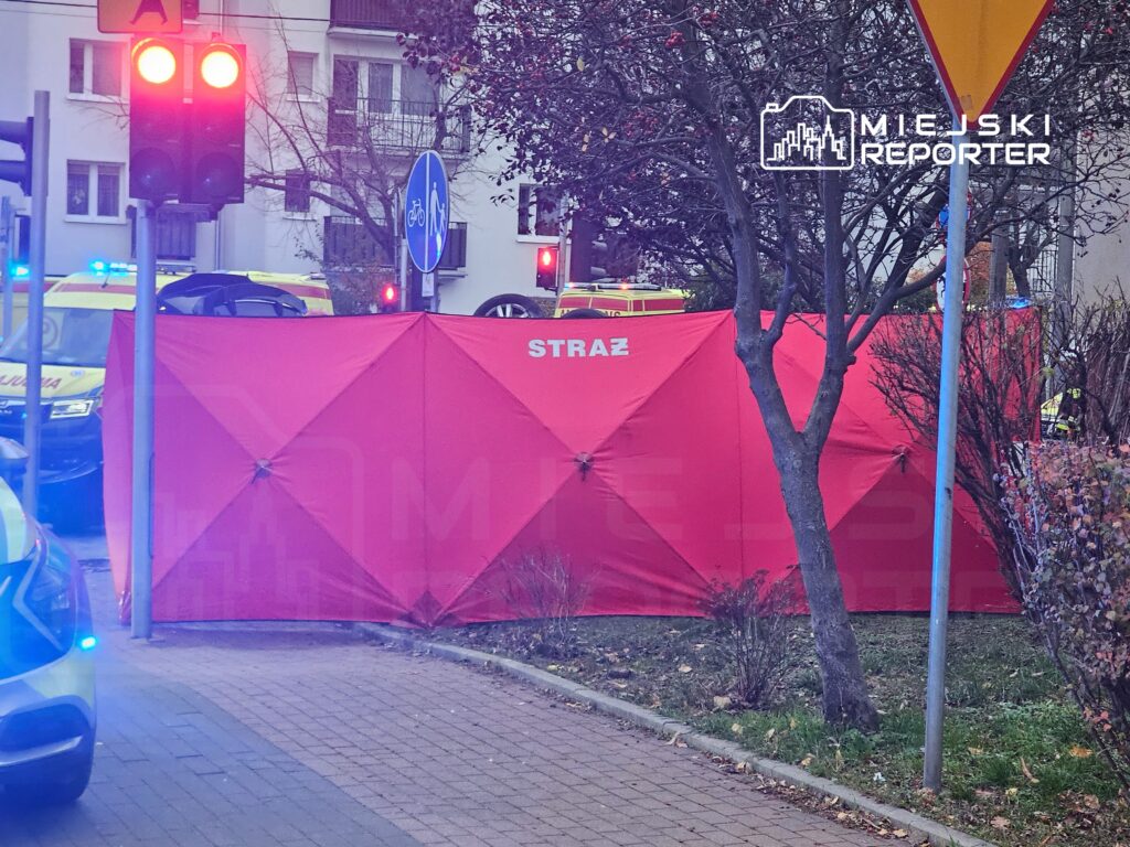 Czerwony namiot strażacki ustawiony na chodniku przy ulicy, w tle widoczne pojazdy służb ratunkowych.