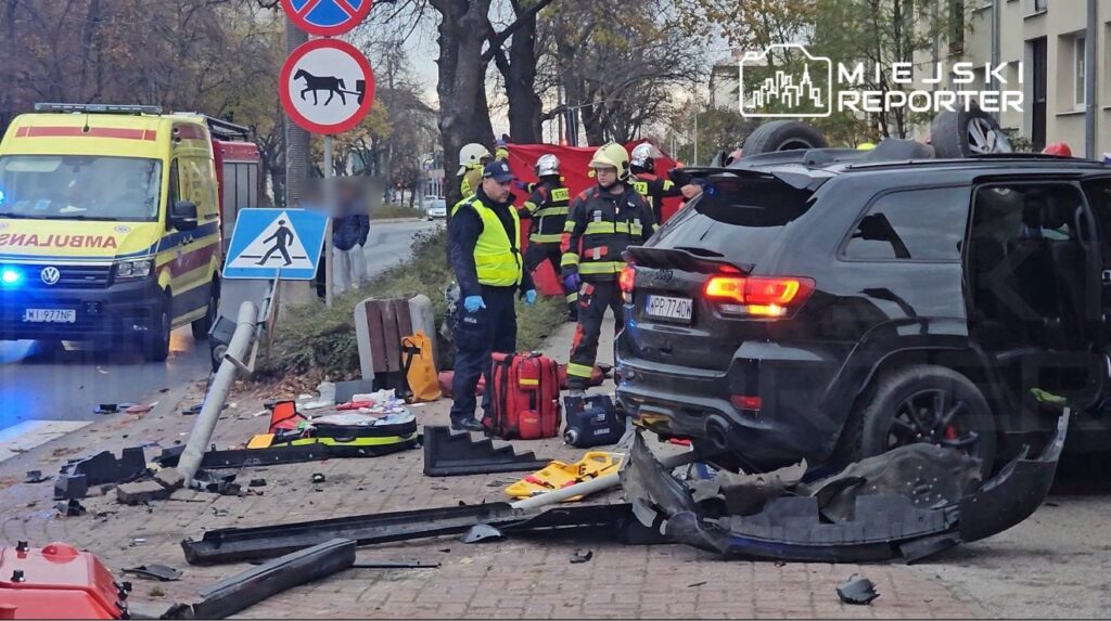 W zderzeniu drogowym uczestniczy czarny samochód osobowy z uszkodzoną tylną częścią, obok stoją ratownicy medyczni i strażacy. W tle widać ambulans oraz znaki drogowe.