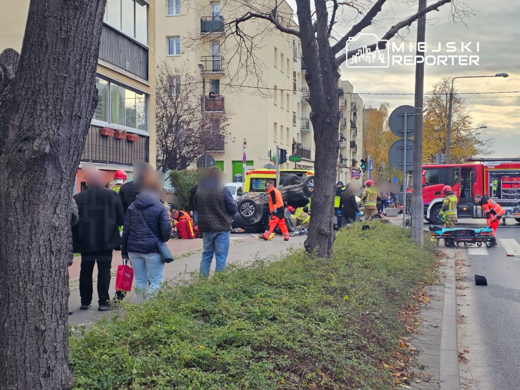 Grupa ratowników i przechodniów zbiera się wokół przewróconego pojazdu na ulicy, obok interweniujących służb.