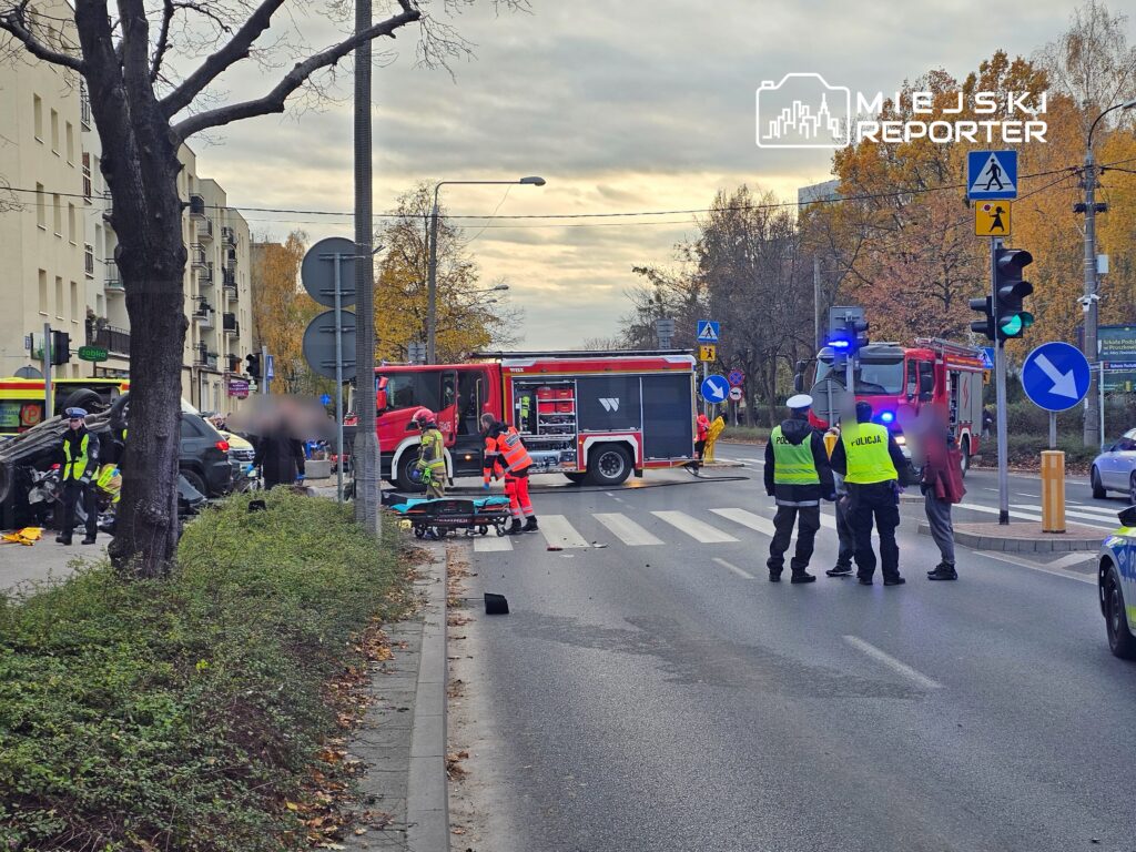 Na skrzyżowaniu ulicy interweniują strażacy i policjanci, zablokowane są pojazdy. W tle widoczny wóz strażacki.