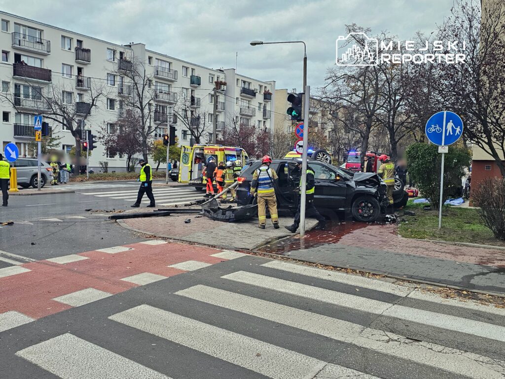 W miejscu wypadku drogowego pracują ratownicy medyczni i strażacy, obok uszkodzonego samochodu na skrzyżowaniu.