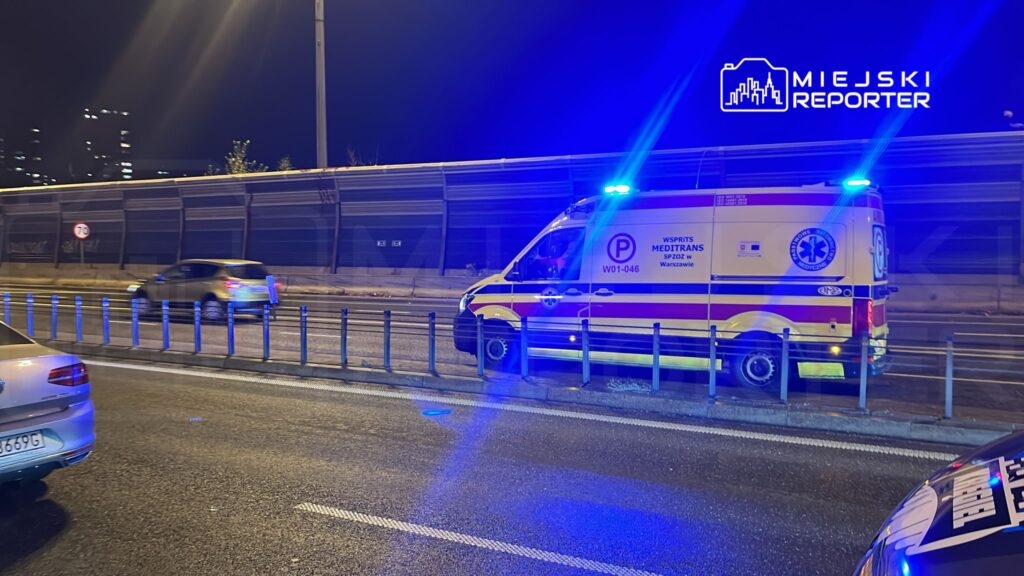 Karetka pogotowia z włączonymi sygnałami świetlnymi zatrzymana na drodze, obok przejeżdżające samochody w nocnej scenerii.