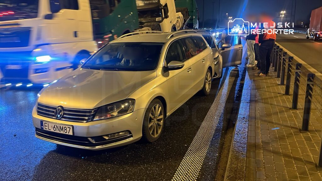 Złoty samochód osobowy stoi na poboczu drogi, obok grupka osób rozmawia, w tle widoczne ciężarówki z oświetleniem.