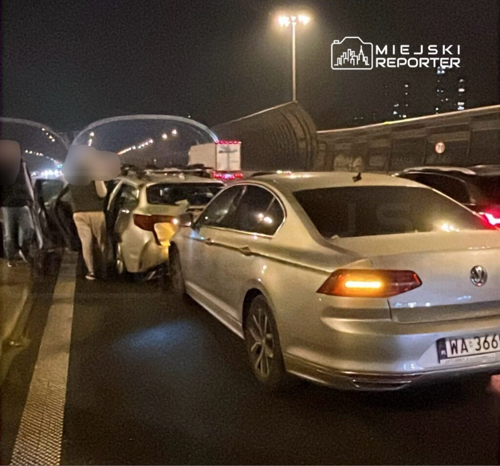 Kilka osób stoi obok uszkodzonego samochodu na drodze, w tle widać inne pojazdy i oświetlenie uliczne.