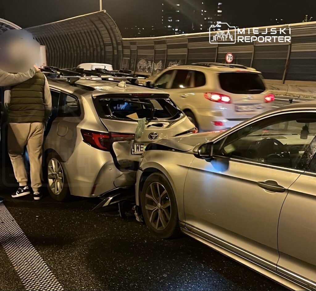 Mężczyzna w odzieży sportowej stoi obok uszkodzonego samochodu na ruchliwej drodze, obok innych pojazdów.