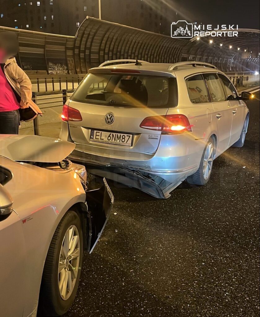 Kobieta w jasnej kurtce stoi obok uszkodzonego samochodu, na drodze w nocy, z widoczną kolizją dwóch pojazdów.