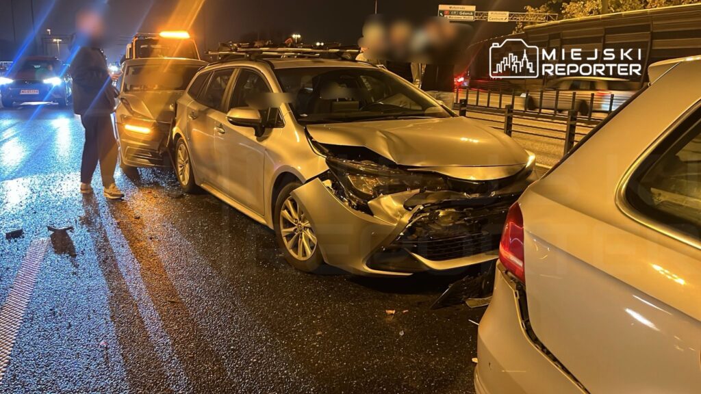 Mężczyzna w dresie stoi obok uszkodzonego samochodu na zatłoczonej drodze nocą, w tle inne pojazdy.