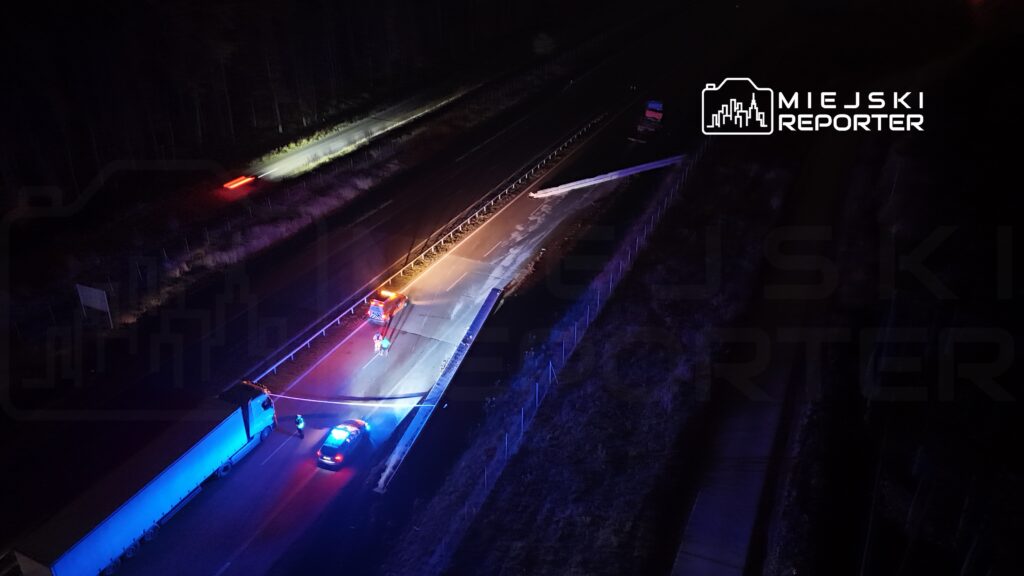 Na drodze leży przewrócony słup, obok policyjne radiowozy i strażacy zabezpieczający miejsce zdarzenia.