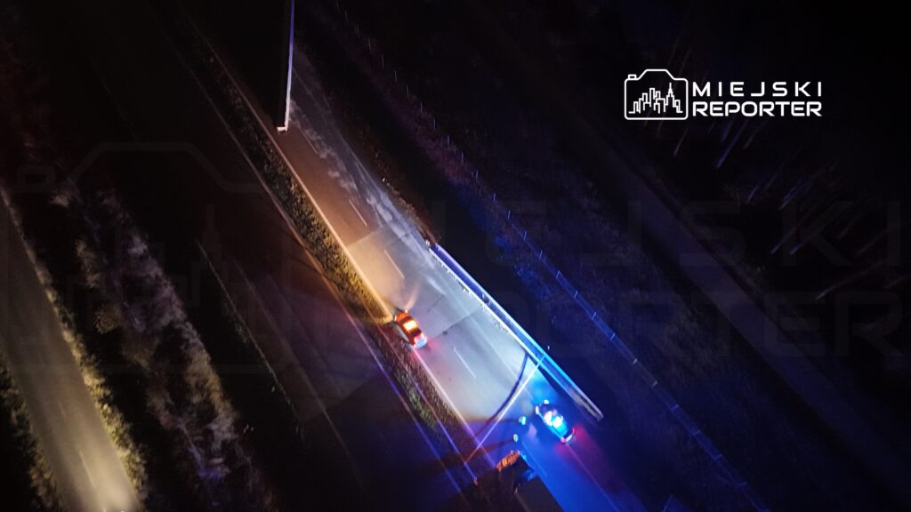 Na drodze nocą zatrzymane są dwa samochody, w tym jeden z włączonymi sygnałami policyjnymi.