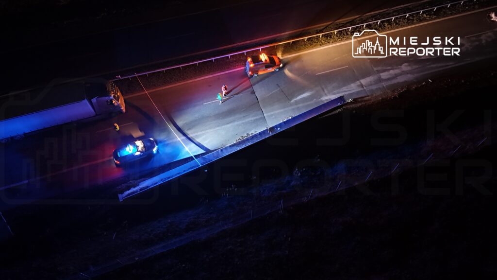 Na drodze stoi policjant z latarką, obok zaparkowane radiowozy z włączonymi sygnałami świetlnymi.